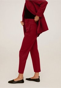 Persoon die donkerrode op maat gemaakte broek en bijpassende blazer aanpast, gekleed in een zwart top en zwarte platte schoenen, staande tegen een effen achtergrond.