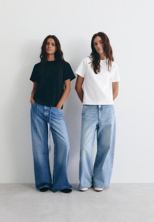 Dve ženski stojita drug ob drugi in nosita široke modre jeans hlače; ena ima črno majico, druga belo, pred enobarvno belo steno.