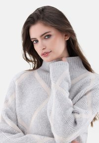 Hellgrauer Pullover mit hohem Kragen, mit strukturiertem Strick und diagonalen beigen Streifen. Weicher Stoff mit locker sitzenden Ärmeln.