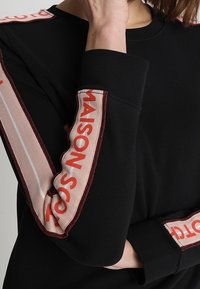 Czarny longsleeve z długim rękawem z kontrastowymi paskami tekstowymi w odcieniach beżu i czerwieni wzdłuż rękawów. Posiada okrągły dekolt i miękką teksturę tkaniny.