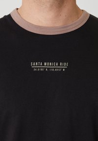 Primer plano de una camiseta negra con cuello marrón, con el texto "SANTA MONICA RIDE" y coordenadas geográficas debajo del texto.