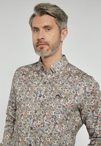 Koszulka z kwiatowym wzorem zapinana na guziki, na beżowym tle z wielokolorowymi kwiatami, z długimi rękawami i kołnierzykiem. Zawiera detal z logo.