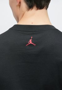 Μαύρο βαμβακερό μπλουζάκι με το κόκκινο λογότυπο Jumpman στην κεντρική θέση πίσω από το λαιμό. Ομαλή υφή, κλασικός σχεδιασμός με στρογγυλή λαιμόκοψη.