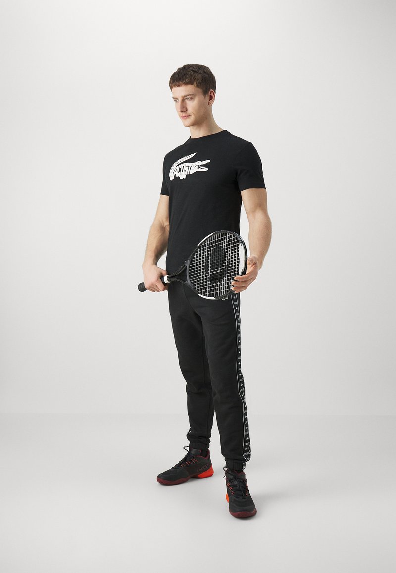 Czarna koszulka z graficznym nadrukiem, czarne dresy z białymi paskami po bokach i czarne buty sportowe z czerwonymi akcentami; trzymając rakietę tenisową.