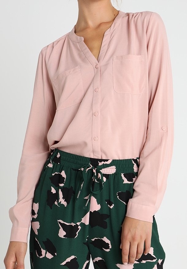 Personne portant un chemisier rose clair à boutons avec deux poches poitrine et un pantalon vert à cordon avec des motifs abstraits noirs et roses.