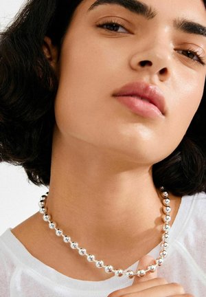 Srebrna ogrlica s perlicama koja se sastoji od glatkih okruglih perli različitih veličina, ležeći na liniji vrata osobe koja nosi bijelu majicu.