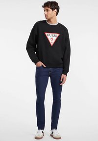 Felpa nera con logo triangolare "GUESS?" rosso e bianco, abbinata a jeans skinny blu scuro e sneakers bianche. Colletto semplice.
