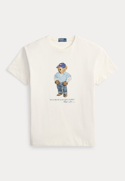Fehér pamut póló, amelyen egy medve grafikája látható, aki kék inget, farmert és kék sapkát visel. Tartalmazza a "Polo Bear by Ralph Lauren" feliratot.