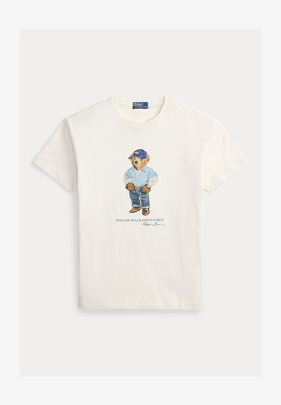 Fehér pamut póló, amelyen egy medve grafikája látható, aki kék inget, farmert és kék sapkát visel. Tartalmazza a "Polo Bear by Ralph Lauren" feliratot.