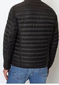 Mand iført en sort quiltet jakke og blå jeans, vist fra ryggen mod en ensfarvet baggrund.