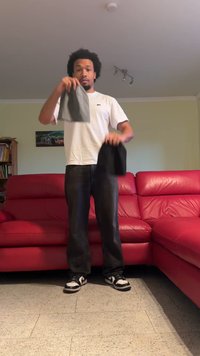 Pelēki un melni cepures, ko tur cilvēks baltā T-kreklā, stāvot uz teksturēta grīdas priekšā sarkanai dīvānam. Spilgta telpu apgaismojums.