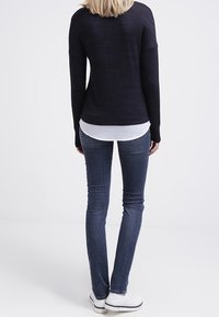 Osoba stojąca tyłem, ubrana w czarną bluzkę z długim rękawem, białą koszulę pod spodem, ciemne skinny jeans i białe buty.