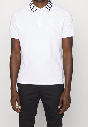 Uomo che indossa una polo bianca con la scritta "JUST" sul colletto e un sottile emblema tonale di tigre sul petto, abbinata a pantaloni neri.