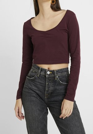 Tummanpunainen pitkähihainen lyhyt yläosa, jossa syvä pyöreä pääntie ja poimittu ommel, yhdistetty korkeavyötäröisiin mustiin farkkuihin, joissa on keltaiset ompeleet.