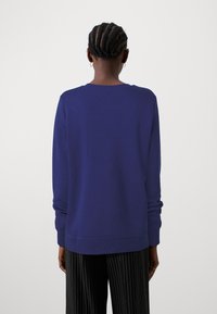 Sudadera morada con cuello redondo, mangas largas y puños acanalados. El material parece suave con una textura lisa; sin patrones visibles.