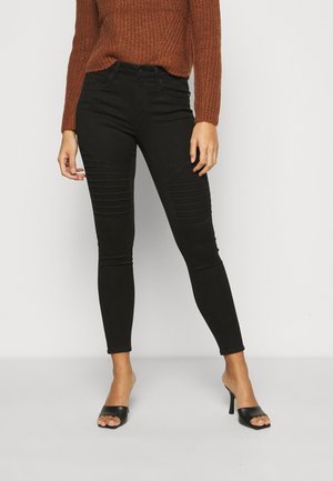 Jeans skinny noirs avec une texture lisse et des détails côtelés sur les cuisses, associés à un pull en tricot de couleur rouille et des sandales à talons noires.