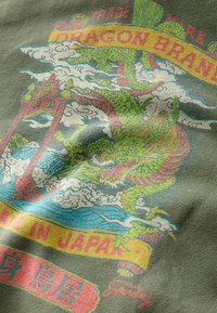 T-shirt verde con una grafica di un drago colorato, nuvole ed elementi di testo. Il design include dettagli in rosso, giallo e blu.