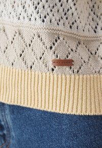 Kreminė megzta palaidinė su deimanto raštais, kreminiu ir geltonu pašonu, o ant rudos odos etiketės užrašyta "PEPE JEANS".