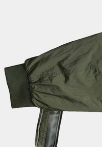 Manica di giacca bomber verde oliva con polsino elastico, dotata di una zip nera lucida e tessuto testurizzato. Costruzione a pannelli con tessuto arricciato.