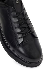 Zwarte leren sneaker met een gladde neuskap, gestikte details, zwarte veters die door vijf paar oogjes zijn geregen.