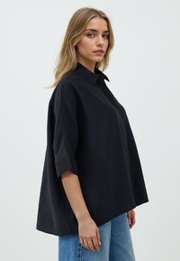 Black, oversized short-sleeve shirt with a collar, made of a smooth fabric. Features a loose fit and subtle stitching details.