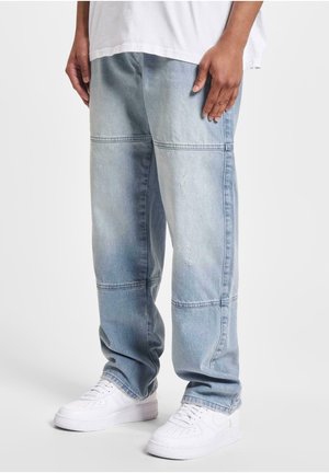 Jeans bleu clair à coupe ample avec des panneaux horizontaux cousus, portés avec des baskets blanches et une chemise blanche sur une personne debout.