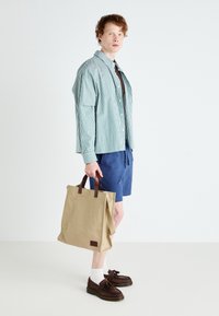 Sac fourre-tout en toile beige avec des poignées en cuir marron. Le modèle porte une chemise à rayures verte à boutons et un short bleu, accompagnés de chaussures marron.