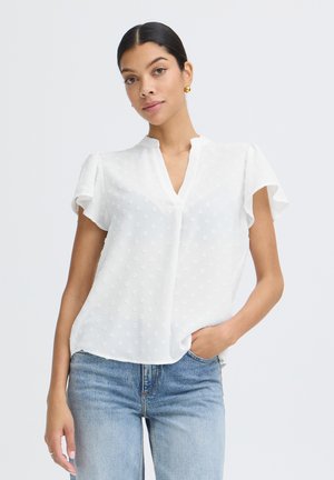 Mladá žena nosí bielu textúrovanú blúzku s krátkym rukávom a svetlomodré rifle, pózuje s jednou rukou v vrecku na jednoduchom pozadí.