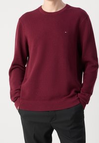 Pull en maille bordeaux avec un motif texturé, col rond, poignets et ourlet côtelés. Présente un petit logo brodé sur la poitrine.