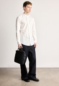 Λευκό πουκάμισο με γιακά και κουμπιά μπροστά, σε συνδυασμό με σκούρο μπλε τζιν φαρδιάς γραμμής και μαύρα παπούτσια, κρατώντας μια μαύρη τσάντα με υφή.