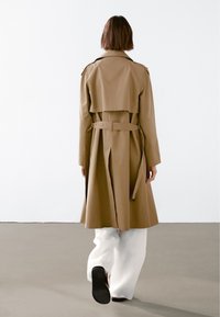 Beżowy płaszcz trench z naramiennikami na ramionach, z paskiem w talii i wentylacją z tyłu. Gładka tekstura, długość do kolan, zestawiony z szerokimi białymi spodniami.