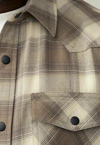 Detailní záběr na béžovo-hnědou kostkovanou košili s černými druky a náprsní kapsou s klopou.