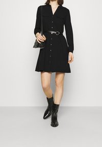 Robe chemise noire à manches longues, deux poches poitrine, taille cintrée par une ceinture, associée à des bottines noires et un sac noir.
