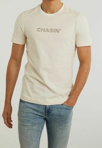 Beige t-paita, jossa on pyöreä kaula-aukko ja lyhyet hihat. Rinnan kohdalla t-paidassa on tummemmasta beigestä painettu "CHASIN'".