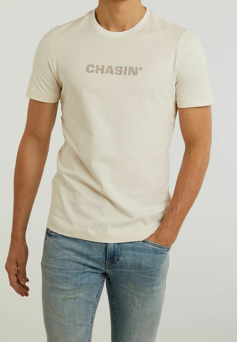 Beige t-paita, jossa on pyöreä kaula-aukko ja lyhyet hihat. Rinnan kohdalla t-paidassa on tummemmasta beigestä painettu "CHASIN'".