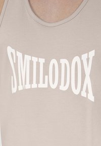 Lichtbeige tanktop met grote witte tekst "SMILODOX" in een vet, gebogen lettertype. De stof lijkt soepel met een eenvoudig ontwerp.