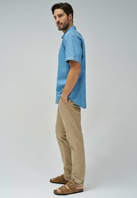 Camisa de manga corta azul claro con cuello abotonado, combinada con pantalones beige y sandalias marrones con dobles correas y hebillas.
