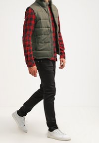 Grüne gepolsterte Weste über einem rot-schwarz karierten Hemd, kombiniert mit schwarzen Jeans und weißen Sneakern. Auffällige Nähte und Designdetails sind sichtbar.