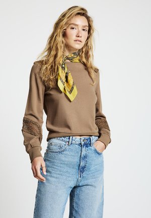 Felpa marrone con dettagli in pizzo sulle maniche, abbinata a jeans a vita alta di colore azzurro chiaro. Completa il look una sciarpa gialla con motivi.