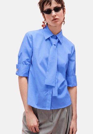 Giovane persona che indossa una camicia a maniche corte blu brillante con cravatta abbinata, pantaloni grigi, occhiali da sole scuri e orecchini arancioni a forma di fiore.