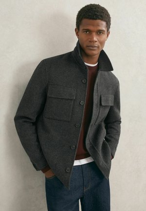 Tmavosivá bunda zo zmesi vlny s konštrukčným golierom, dvoma vreckami vpredu a zapínaním na gombíky. Nosená cez bordový sveter a modré rifle.