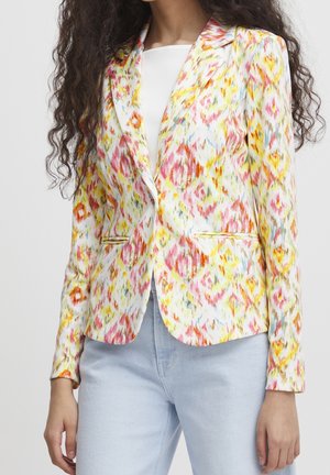 Femme portant un blazer ajusté à motifs multicolores avec des teintes jaunes, roses et orange, par-dessus un haut blanc et un jean bleu clair.