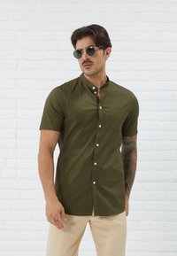 Short-sleeve olive green shirt with a stand collar, front button closure, and a chest pocket, worn with cream-colored pants.