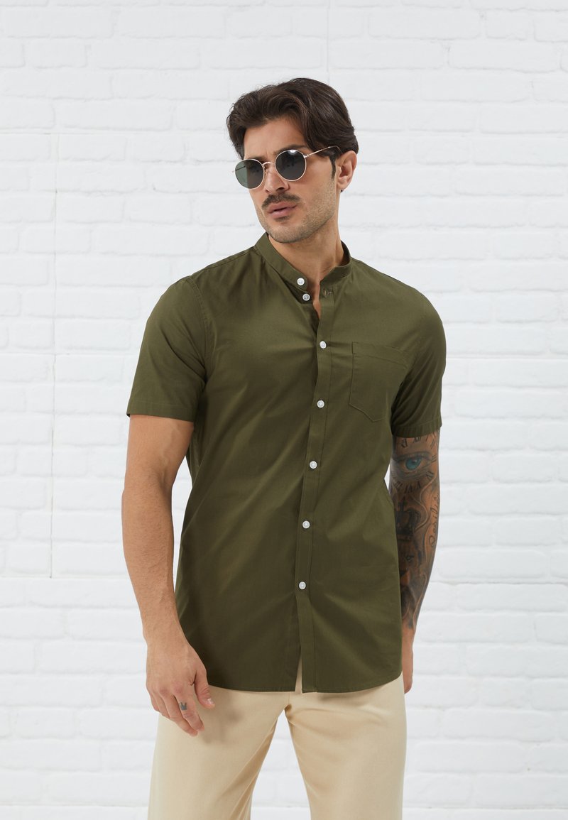 Short-sleeve olive green shirt with a stand collar, front button closure, and a chest pocket, worn with cream-colored pants.