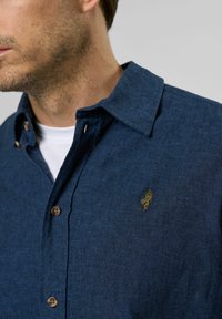 Tummansinisestä painettavasta kankaasta valmistettu nappipaita, jossa on huomaamaton kultainen logo vasemmassa rinnassa ja ruskeat napit.
