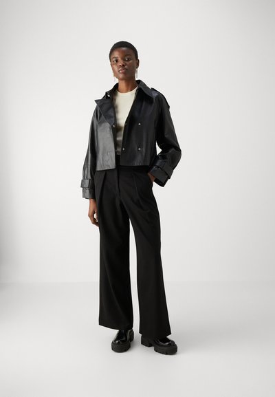 Μαύρο δερμάτινο κομμένο μπουφάν με κουμπιά, φαρδιά πέτα και σφραγίδες στα μανίκια, σε συνδυασμό με ψηλόμεσα μαύρα παντελόνια και χοντρά μαύρα παπούτσια.