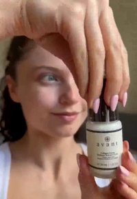 Transparent glasflaska med vit kräm inuti, svart pump-top och svart etikett. Hållen av en hand med långa rosa naglar.