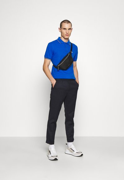 Μπλε πόλο μπλούζα με λευκές λεπτομέρειες, μαύρη τσάντα crossbody, ναυτικές παντελόνες και ελαφριά αθλητικά παπούτσια με μαύρα και λευκά στοιχεία. Λείος υφή.