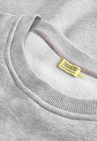 Grauer Sweatshirt mit geripptem Rundhalsausschnitt und weichem Innenfutter. Enthält ein gelbes Etikett mit dem Logo "PEANUTS x CODELLO".