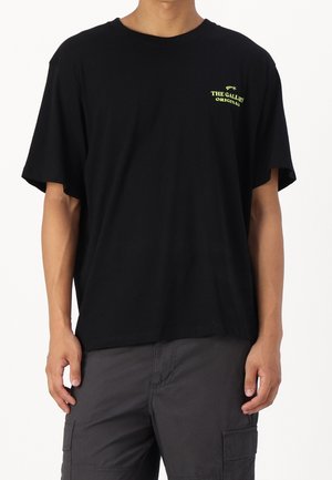 Man draagt een zwart T-shirt met kleine gele tekst "THE GALLERY ORIGINAL" op de linkerbovenborst en donkergrijze cargobroeken, staat met ontspannen armen.